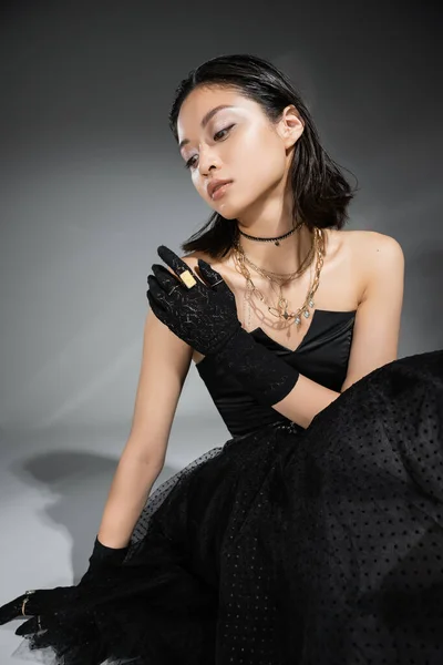 Encantador asiático jovem mulher com cabelo curto sentado em preto vestido sem alças com saia de tule e luvas enquanto olha para o fundo cinza, penteado molhado, colares dourados e anéis — Fotografia de Stock