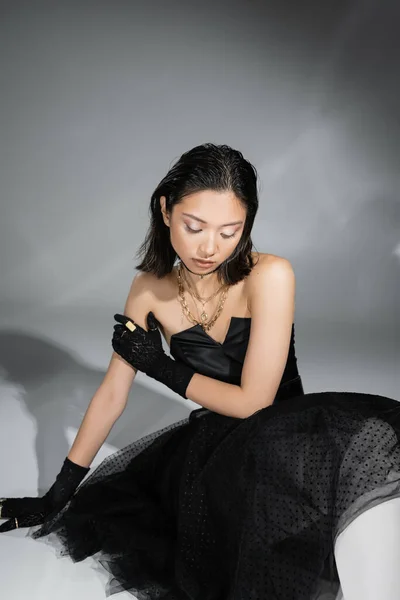 Stunning asian young woman with short hair sitting in black strapless dress with tulle skirt and gloves while looking down on grey background, wet hairstyle, golden necklaces and rings — Stock Photo