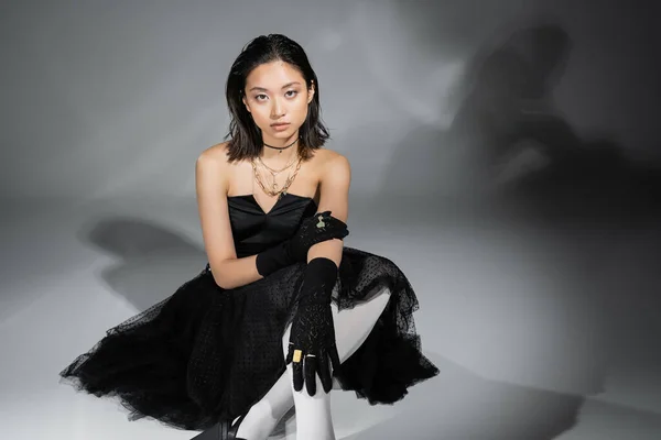 Стильная азиатская молодая женщина с короткими волосами, сидящая в черном платье без бретелек с тюлем юбка, белые колготки и перчатки, глядя на камеру на сером фоне, влажная прическа, золотые ожерелья — стоковое фото