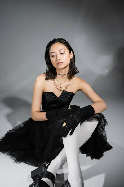 Мечтательная азиатская молодая женщина с короткими волосами сидя в черном платье без бретелек с тюлем юбка белые колготки, обувь и перчатки, глядя в сторону на сером фоне, влажная прическа, золотые ожерелья — стоковое фото