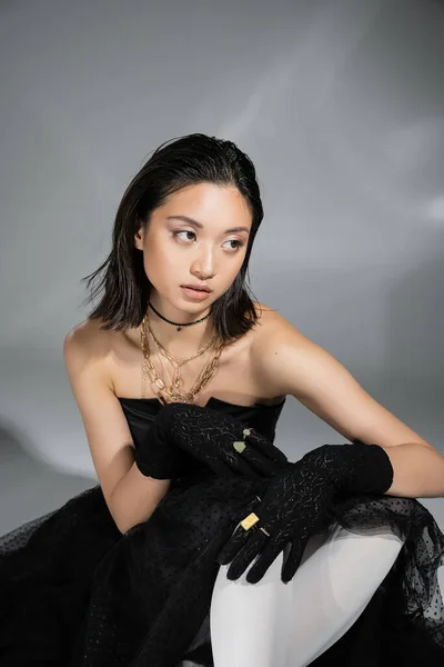 Atractiva mujer joven asiática con pelo corto sentado en vestido negro sin tirantes con falda de tul y guantes mirando hacia otro lado sobre fondo gris, peinado mojado, collares dorados, modelo de ensueño - foto de stock