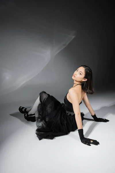 Полная длина азиатская молодая женщина с короткими волосами сидя в черном платье без бретелек с тюлем юбка белые колготки, обувь и перчатки при взгляде на камеру на сером фоне, влажная прическа, увлекательный — стоковое фото