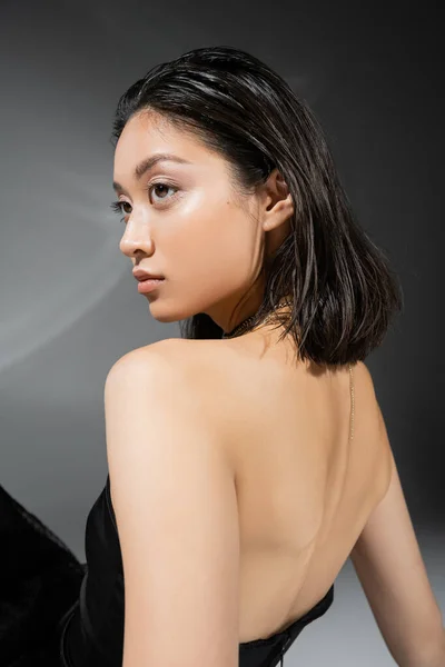 Porträt einer asiatischen jungen Frau mit kurzen brünetten Haaren, die in einem schwarzen schulterfreien Kleid auf grauem Hintergrund steht, Alltagsschminke, Nassfrisur, charmantes Modell, natürliche Schönheit — Stockfoto