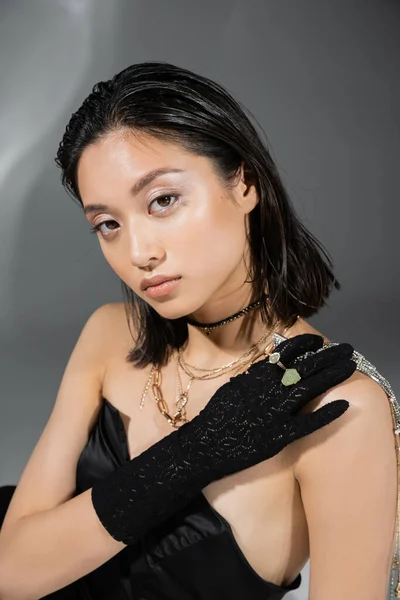 Чарівна азіатська модель з коротким та брюнетковим волоссям, що тримає золоті прикраси в руці, позуючи в плащі та чорну рукавичку на сірому фоні, повсякденний макіяж, мокра зачіска, молода жінка — стокове фото