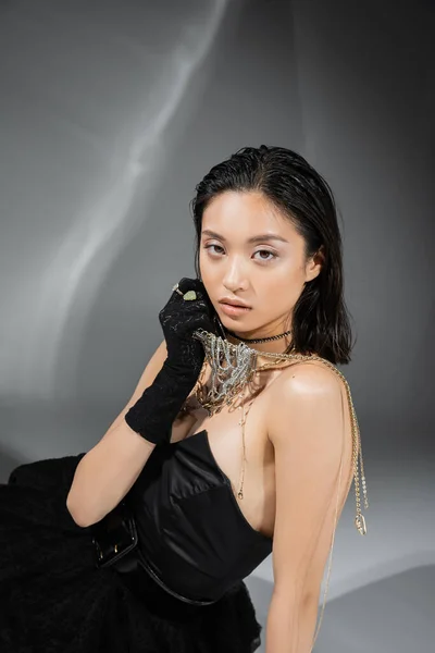 Modelo asiático con pelo corto y morena sosteniendo joyas de oro en la mano mientras posando en vestido sin tirantes y guante negro sobre fondo gris, maquillaje diario, peinado húmedo, mujer joven, glamour — Stock Photo