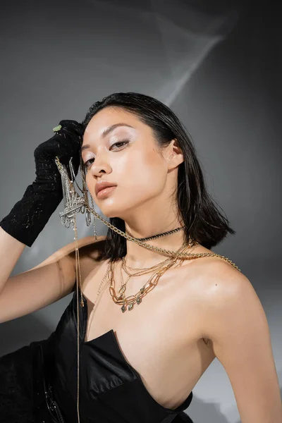 Stunning asian model with short and brunette hair holding golden jewelry in hand while posing in strapless dress and black glove on grey background, everyday makeup, wet hairstyle, young woman — Stock Photo