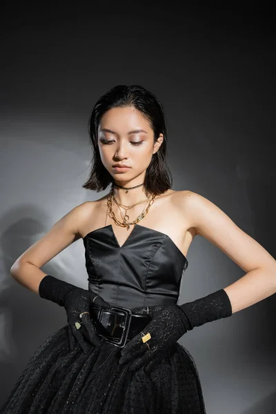 Encantador asiático jovem mulher com cabelo curto posando em preto vestido sem alças com saia de tule ao tocar cinto e olhando para baixo no fundo cinza, penteado molhado, jóias douradas — Fotografia de Stock