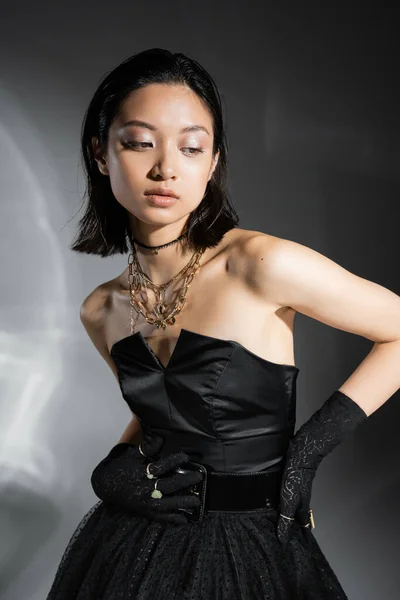 Encantador asiático jovem mulher com cabelo curto posando em vestido sem alças preto com saia de tule com cinto e luvas enquanto olha para o fundo cinza, penteado molhado, jóias douradas — Fotografia de Stock