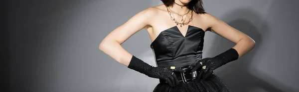Vista recortada de la joven posando en vestido negro sin tirantes con falda de tul y guantes tocando el cinturón y de pie sobre fondo gris, joyas de oro, pancarta - foto de stock