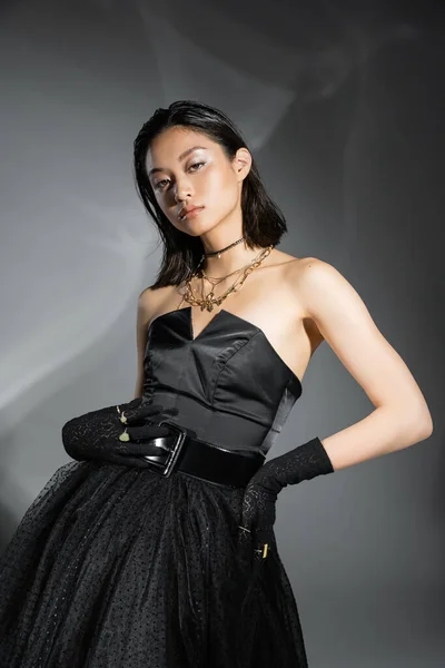 Очаровательная азиатская молодая женщина с короткими волосами позирует в черном платье без бретелек с юбкой тюля с поясом и перчатками, стоя с рукой на бедре на сером фоне, влажная прическа, золотые украшения — стоковое фото