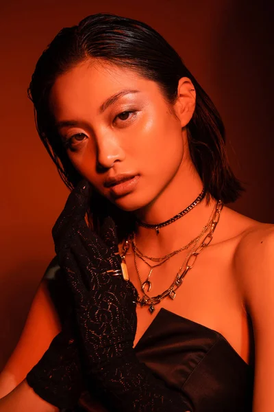 Retrato de sedutor asiático jovem mulher com cabelo curto e penteado molhado posando em preto strapless vestido e luvas enquanto em pé sobre laranja fundo com iluminação vermelha, jóias douradas — Fotografia de Stock