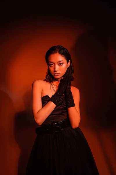 Красивая азиатская женщина с короткими волосами и влажной прической позирует в черном платье без бретелек с тюлем юбка и перчатки, стоя на темно-оранжевом фоне с красным освещением, золотые украшения, молодая модель — стоковое фото