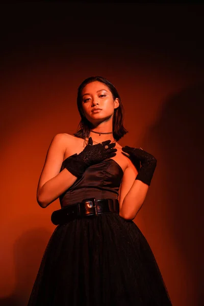 Captivating asian woman with short hair and wet hairstyle posing in black strapless dress with tulle skirt and gloves while standing on orange background with red lighting, golden jewelry, young model — Stock Photo