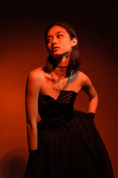Asian woman with short hair and wet hairstyle posing with hand on hip in black strapless dress with tulle skirt and gloves while standing on orange background with red lighting, golden jewelry — Stock Photo