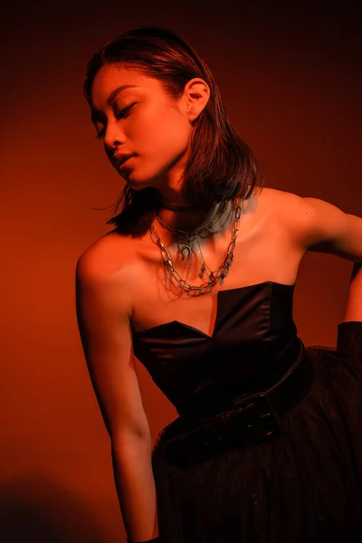 Hipnotizante mulher asiática com cabelo curto e penteado molhado posando em vestido sem alças preto com saia de tule e luvas enquanto em pé sobre fundo laranja com iluminação vermelha, jóias douradas, jovem modelo — Fotografia de Stock
