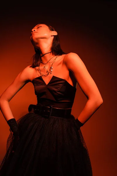 Mujer asiática elegante con los ojos cerrados y el peinado mojado posando en vestido negro sin tirantes con falda de tul y guantes, mientras que de pie sobre fondo naranja con iluminación roja, joyas de oro, modelo joven - foto de stock