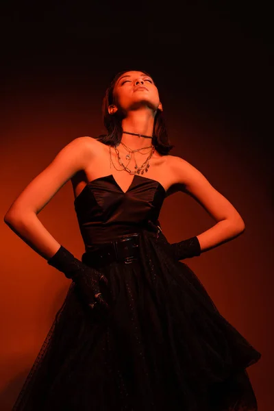 Stylish asian woman with closed eyes and wet hairstyle posing with hands on hips in black strapless dress with tulle skirt and gloves while standing on orange background with red lighting, young model — Stock Photo