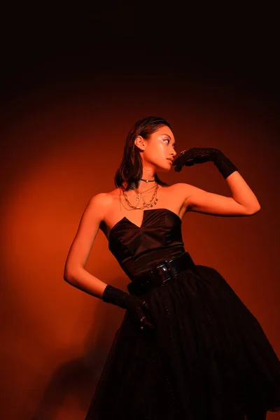 Сияющая азиатская женщина с короткими волосами и влажной прической, позирующая с рукой на бедре в черном платье без бретелек с юбкой тюля и перчатками, стоя на оранжевом фоне с красным освещением, молодая модель — стоковое фото