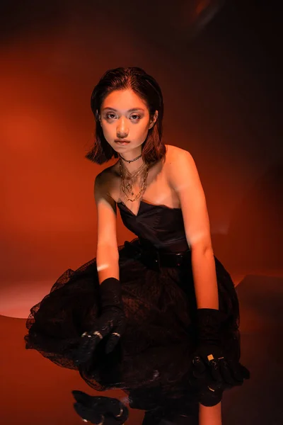 Asian woman with short hair and wet hairstyle posing in stylish black strapless dress with tulle skirt and gloves while standing on orange background with red lighting, golden jewelry, young model — Stock Photo