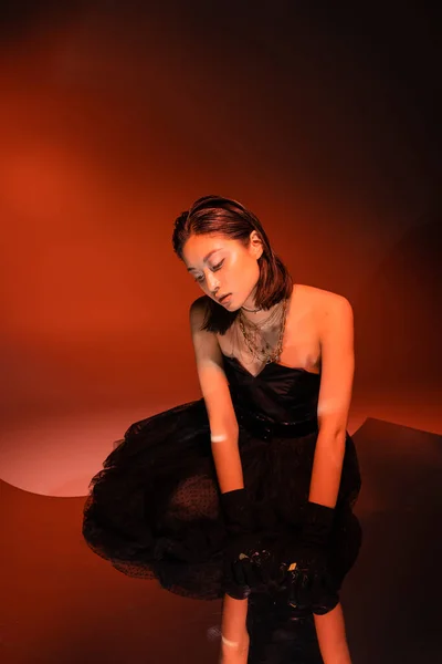 Заманчивая азиатская женщина с короткими волосами и влажной прической позирует в стильном платье без бретелек с тюлем юбка и перчатки, стоя на оранжевом фоне с красным освещением, золотые украшения, молодая модель — стоковое фото