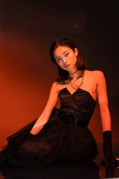 Beautiful asian woman with short hair and wet hairstyle posing in strapless dress with tulle skirt and gloves while standing on orange background with red lighting, young model, looking at camera — Stock Photo