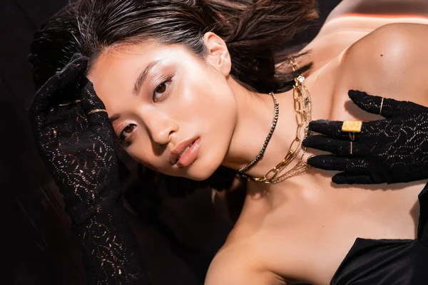 Верхний вид красивой азиатской женщины с короткими волосами и влажной прической позируя в черных перчатках с золотыми кольцами, глядя на камеру на темном фоне, молодой модели — стоковое фото