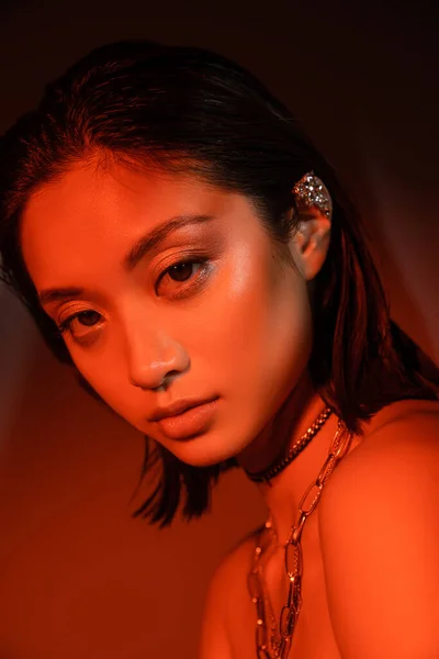Портрет завораживающей азиатской женщины с короткими волосами и влажной прической, позирующей с модной сережкой манжеты и ожерельями на темно-оранжевом фоне с красным освещением, молодой моделью, смотрящей в камеру — стоковое фото