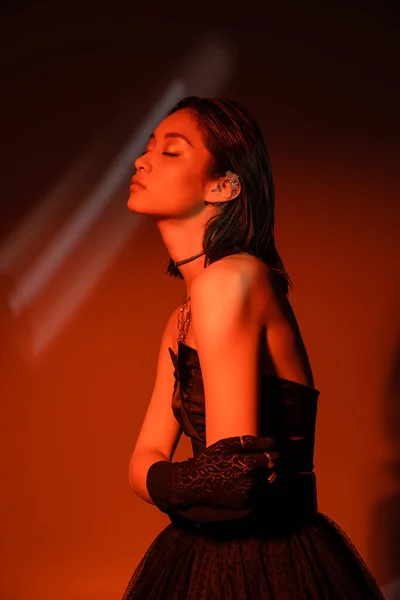 Portrait of stunning asian woman with short hair and wet hairstyle posing in strapless dress and glove with trendy cuff earring and necklaces on dark orange background with red lighting, young model — Stock Photo