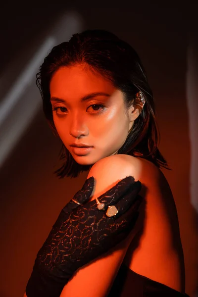Retrato de encantadora mulher asiática com cabelo curto e penteado molhado posando em luva preta com anéis dourados e olhando para a câmera no fundo escuro com iluminação vermelha, modelo jovem, brinco de punho — Fotografia de Stock
