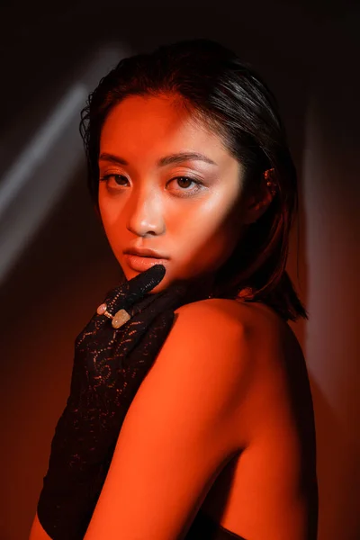 Retrato de jovem mulher asiática com cabelo curto e penteado molhado posando em luva preta com anéis dourados e olhando para a câmera no fundo escuro com iluminação vermelha, modelo, brinco de punho — Fotografia de Stock