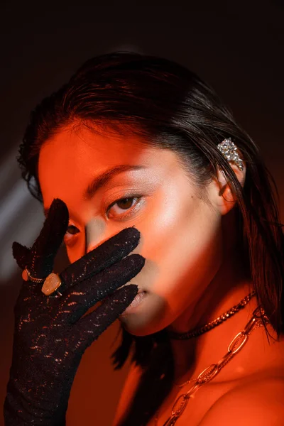 Portrait of alluring asian woman with short hair and wet hairstyle posing in black glove with golden rings and covering face while looking at camera on dark background with red lighting, cuff earring — Stock Photo