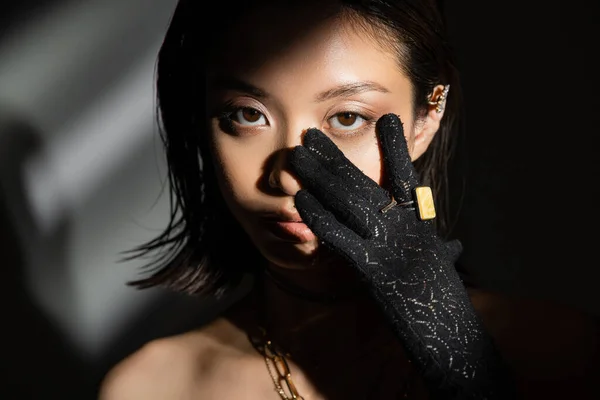 Portrait de jeune femme asiatique à la coiffure mouillée et aux cheveux courts en gant noir avec des anneaux dorés touchant le visage tout en se tenant sur fond gris, mannequin, regardant caméra, ombres, sombre — Photo de stock