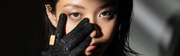 Portrait of asian young woman with wet hairstyle and short hair in black glove with golden rings touching face while standing on grey background, model, looking at camera, shadows, dark, banner — Stock Photo