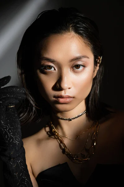 Portrait d'une jeune femme asiatique enchanteresse à la coiffure mouillée et aux cheveux courts posant en gant noir debout sur fond gris, mannequin, regardant caméra, ombres, fard à paupières sombre et chatoyant — Photo de stock