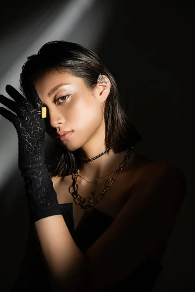 Portrait de jeune femme asiatique enchanteresse à la coiffure mouillée et aux cheveux courts posant en gant noir avec anneaux dorés et manchette d'oreille debout sur fond gris, jeune mannequin, ombres, sombre — Photo de stock