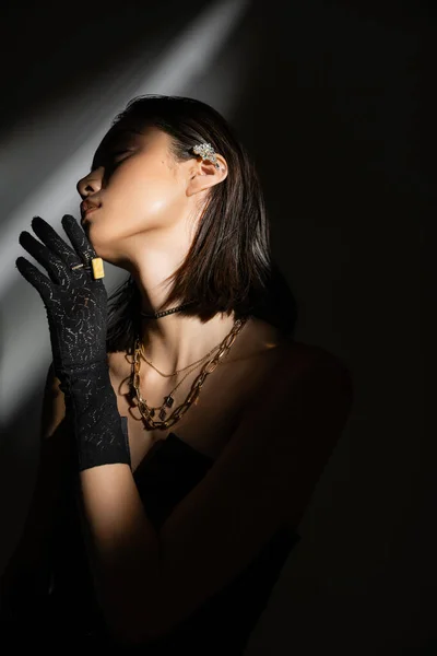 Captivating asian young woman with wet hairstyle and short hair posing in strapless dress and black glove with rings while standing on grey background, young model, looking away, shadows, dark — Stock Photo