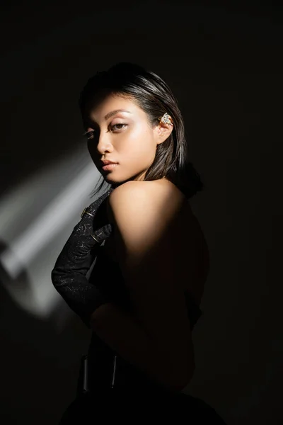 Élégante jeune femme asiatique avec coiffure mouillée et cheveux courts posant en robe bustier et gant noir debout sur fond gris, jeune modèle, regardant caméra, ombres, sombre — Photo de stock