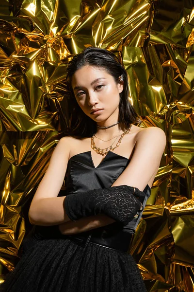 Elegante asiatische junge Frau mit nasser Frisur und kurzen Haaren posiert mit verschränkten Armen im schwarzen trägerlosen Kleid mit Handschuhen neben glänzendem Hintergrund, faltige Goldfolie — Stockfoto