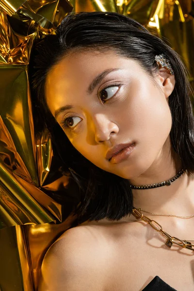 Portrait de séduisante jeune femme asiatique aux cheveux courts mouillés posant à côté de fond jaune brillant, mannequin, détournant les yeux, feuille d'or ridée, beauté asiatique, maquillage naturel — Photo de stock