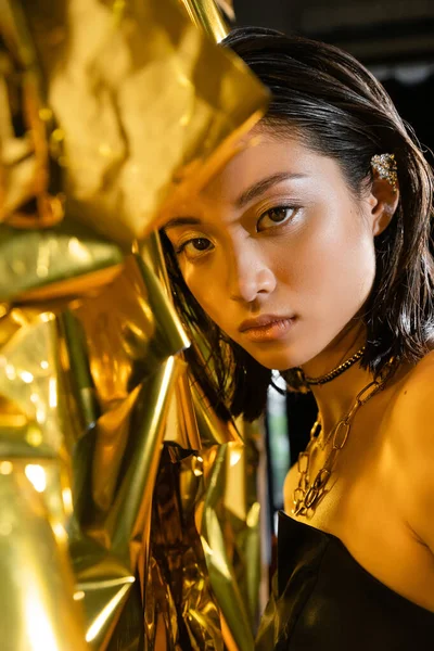 Porträt von glamourösen asiatischen junge Frau mit nassen kurzen Haaren posiert in trägerlosen Kleid neben glänzenden Hintergrund, Modell, Blick in die Kamera, faltige Goldfolie, natürliche asiatische Schönheit — Stockfoto