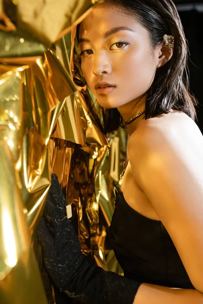 Retrato de graciosa asiática jovem mulher com cabelo curto molhado posando em vestido preto sem alças ao lado de fundo amarelo brilhante, modelo, olhando para a câmera, enrugado folha dourada, natural asiático beleza — Fotografia de Stock