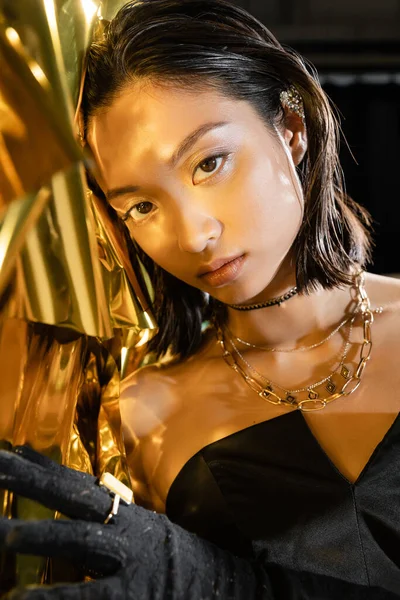 Porträt einer schicken asiatischen jungen Frau mit nassen kurzen Haaren, die in einem schwarzen schulterfreien Kleid vor glänzendem Hintergrund posiert, Modell, in die Kamera schauend, faltige Goldfolie, natürliche asiatische Schönheit — Stockfoto