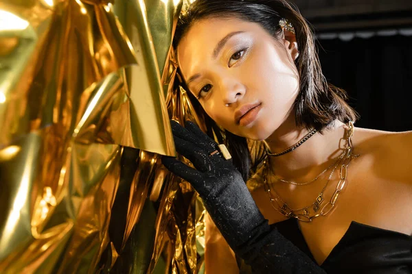 Portrait de séduisante jeune femme asiatique aux cheveux courts mouillés et au gant noir touchant fond jaune brillant, mannequin, regardant la caméra, feuille d'or ridée, beauté asiatique naturelle — Photo de stock