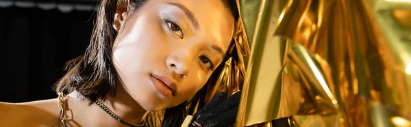 Retrato de seductora mujer joven asiática con el pelo corto mojado y joyas posando junto a fondo dorado brillante, modelo, mirando a la cámara, lámina arrugada, belleza asiática natural, pancarta - foto de stock