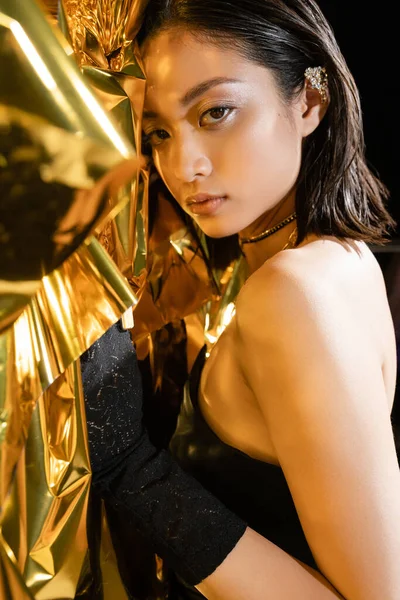 Portrait of alluring asian young woman with wet short hair posing in strapless dress next to shiny background, model, looking at camera, wrinkled golden foil, natural asian beauty — Stock Photo
