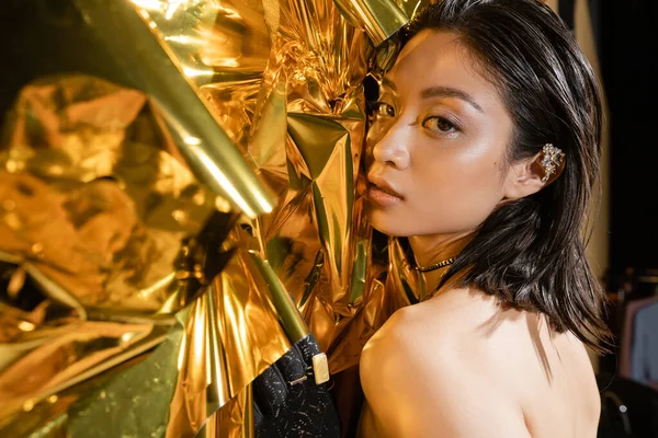 Portrait de séduisante jeune femme asiatique aux cheveux courts mouillés posant à côté d'un fond doré brillant, mannequin, regardant la caméra, feuille jaune ridée, beauté naturelle, gant noir — Photo de stock