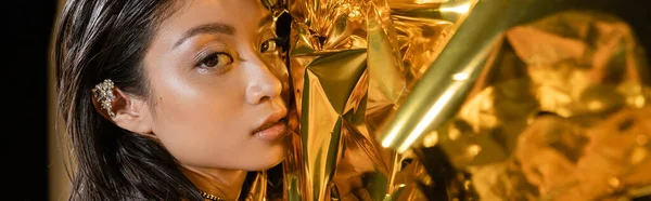 Retrato de seductora mujer joven asiática con el pelo corto húmedo y el manguito de la oreja posando junto al fondo amarillo brillante, modelo, mirando a la cámara, lámina dorada arrugada, belleza natural, pancarta - foto de stock