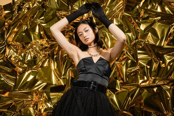 Asian young woman with wet hairstyle and short hair posing in black strapless dress with gloves while standing with raised hands next to shiny yellow background, looking at camera, golden foil — Stock Photo