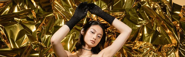 Asiatique jeune femme avec la coiffure humide et les cheveux courts posant dans des gants noirs tout en se tenant avec les mains levées à côté de fond brillant, en regardant la caméra, ridée feuille d'or, bannière — Photo de stock