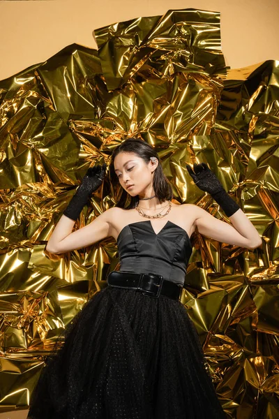 Elegant asian young woman with wet hairstyle and short hair posing in black strapless dress with tulle skirt and gloves while standing next to shiny background, model, wrinkled golden foil — Stock Photo